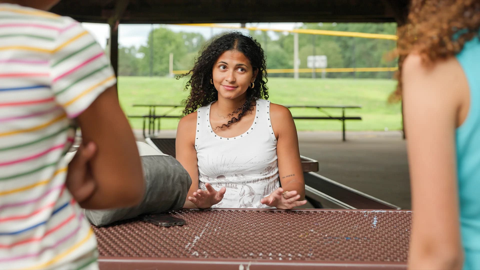 A young woman smiles at two friends across a picnic table under a park pavilion in Along the Way